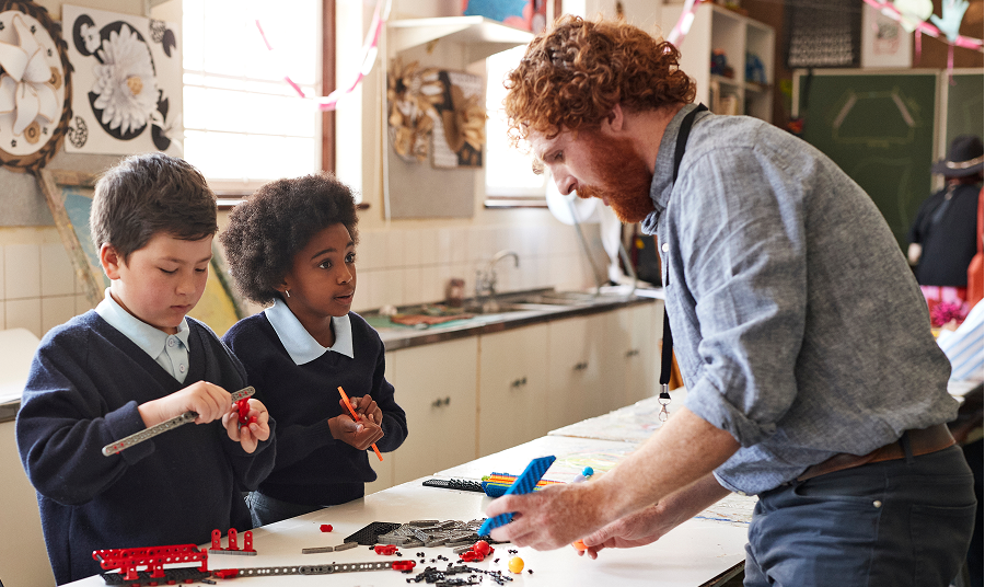 A teacher actively engaging with two young students who are building and experimenting with educational toys or robotics in a classroom.